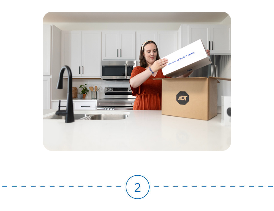An ADT technician uses a power drill to install a security device outside a house while standing on a ladder.