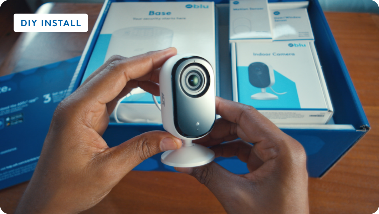 Hands holding a white indoor security camera over an open box of Blue by ADT components, labeled DIY Install.