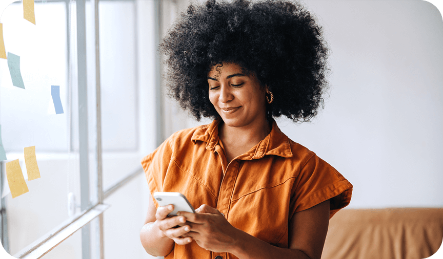 Smiling woman with curly hair in an orange shirt using her smartphone near a window.